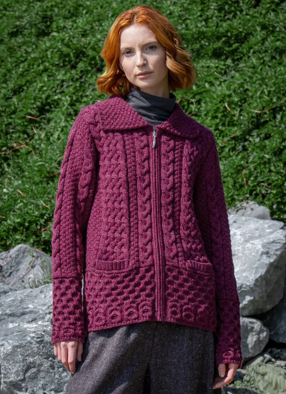 Red head model in front of greenery in grace full zip aran cardigan in raspberry melange